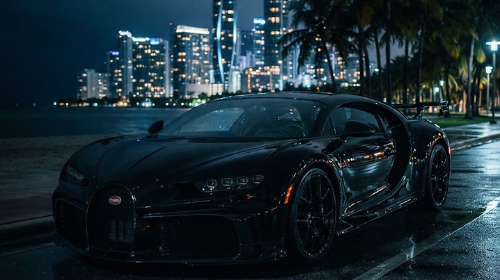 A black Bugatti supercar parked at night on a wet oceanfront boulevard in Miami, palm trees and high-rises reflected in its polished paint
