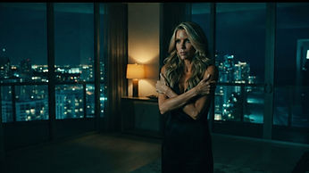 Paige stands tense and alone at night at the floor-to-ceiling windows of her dark waterfront condo, the Miami skyline glowing beyond