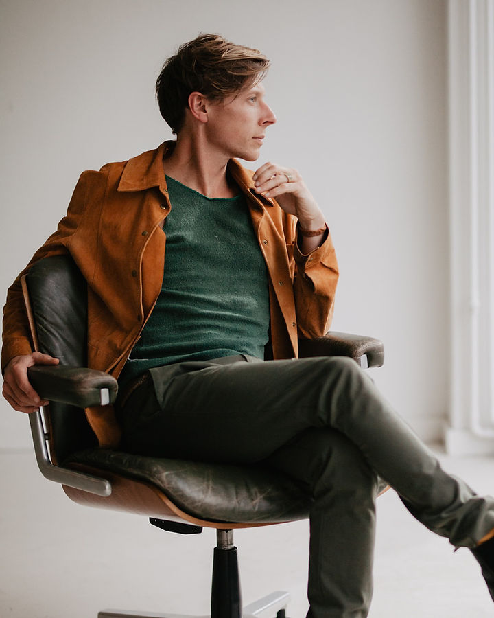 Portrait of Sebastiaan Gamelkoorn, a multi-disciplinary creative based in the Netherlands, seated in a mid-century office chair wearing a suede jacket and green tank top for his professional biography