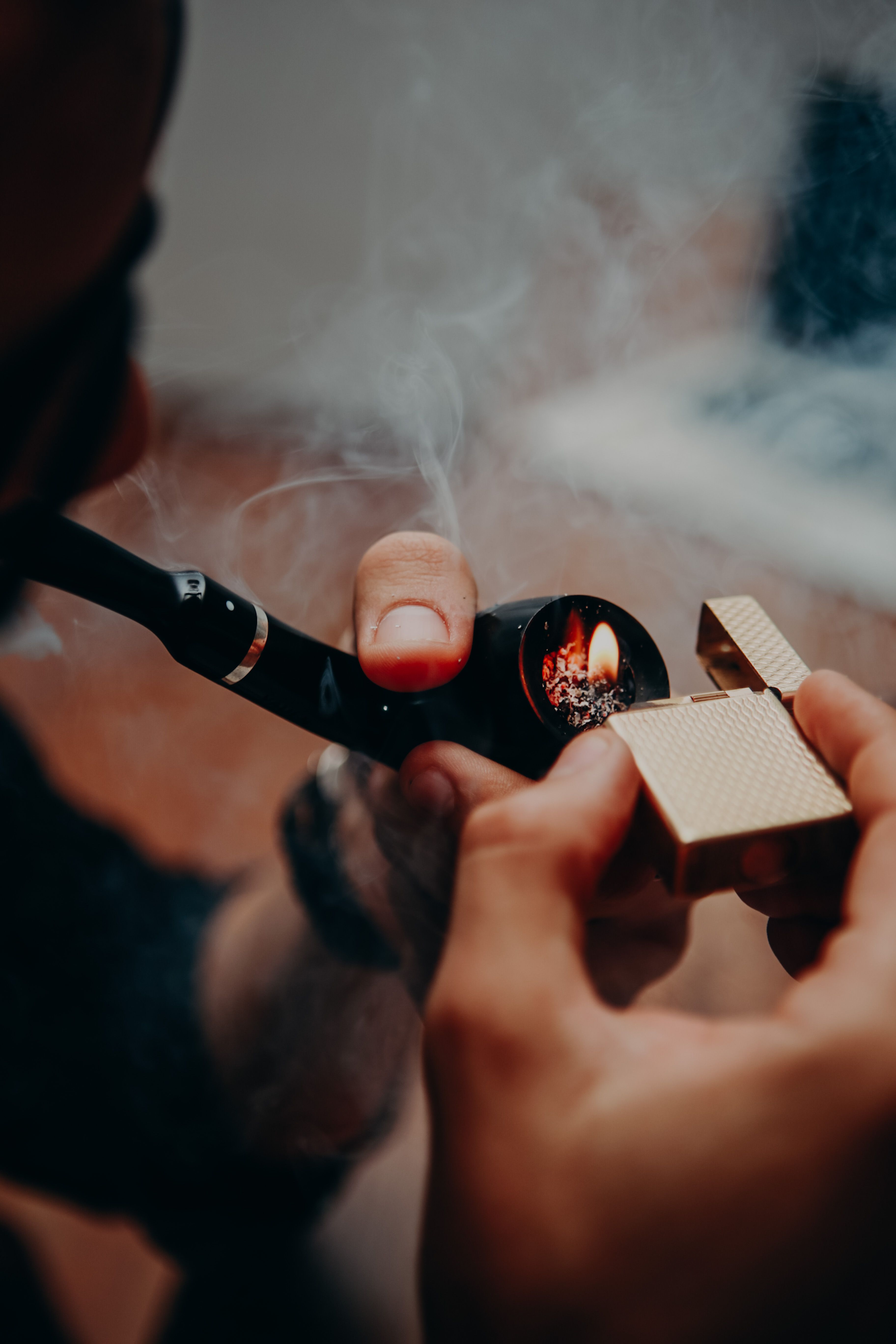 A hand lighting a pipe with a gold lighter, an ember glowing in the bowl and smoke curling upward