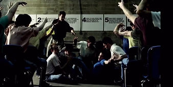 Group footage in a cinder-block room: clients seated in a ring around someone on the floor, the numbered steps posted above them.