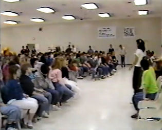 A wide shot of a meeting: rows of participants facing a speaker under a grid of fluorescent lights.