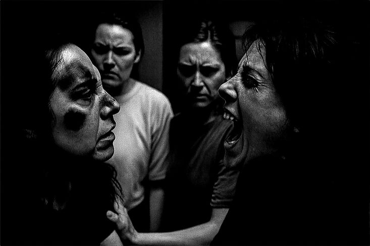 Two clients in profile, one shouting into the other’s face, the group watching from the dark behind them