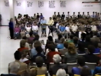 An open meeting filmed from the back of the room: a large group seated in a U-shape, rules posted along the wall.