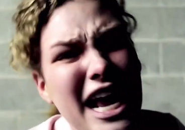 A close-up of a young woman with curly hair mid-scream, harsh light across her face, against a concrete block wall