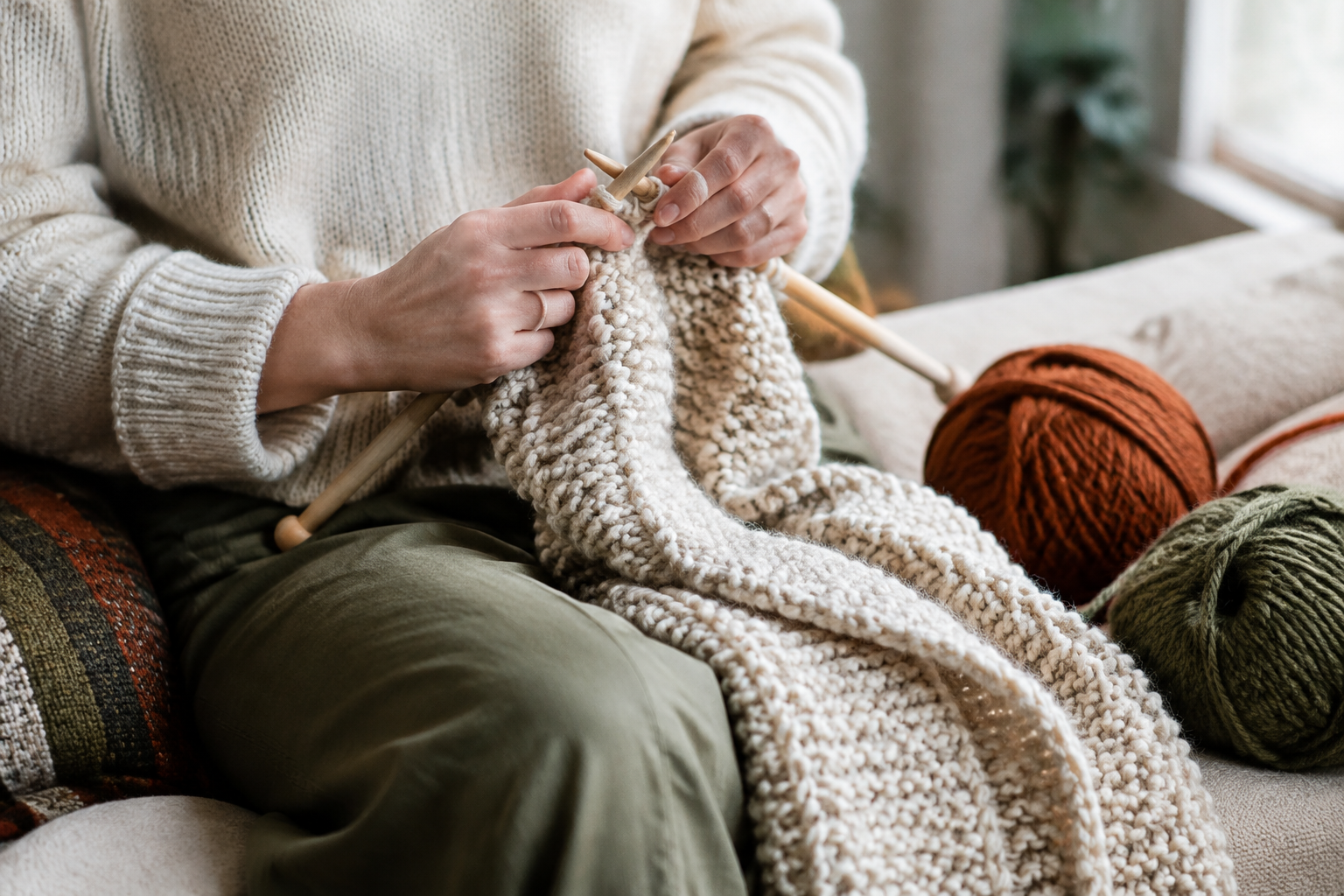 Hands knitting a chunky cream wool scarf with wooden needles and yarn balls