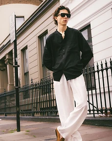 Male model in a black shirt and white trousers standing against a white building with iron railings
