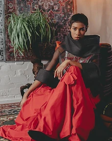 Model in a bright red skirt and black sheer top seated on an upholstered chair against a patterned rug