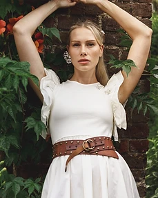 Model in a white top and studded leather belt posing with arms raised against a vine-covered brick wall