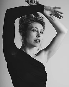 Monochrome portrait of a woman with arms raised overhead in a black one-shoulder top against a grey backdrop