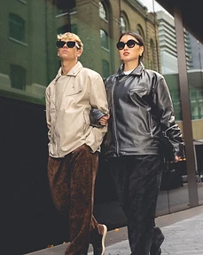 Two models in leather jackets walking arm in arm along a city street beside a glass facade