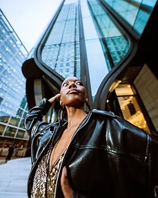 Model in braids wearing a gold sequin top and black leather jacket beneath modern glass architecture