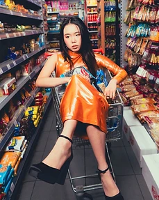 Model in a metallic orange latex dress seated in a shopping cart in a supermarket aisle