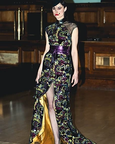 Runway model in a black dress with neon embroidery and a purple sash walking the catwalk