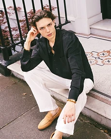 Male model in a black shirt and white trousers squatting on a stone step before a pillar