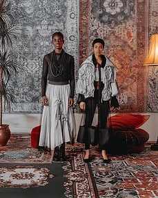 Two models in layered editorial outfits standing before a hanging tapestry in a studio