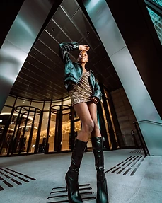 Full-body low-angle shot of a model in a gold sequin dress and leather jacket posing before glass doors at night