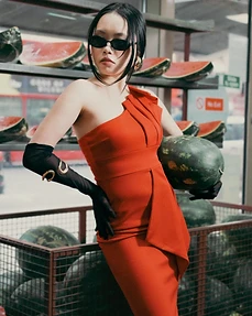 Model in a red one-shoulder dress and black gloves holding a watermelon in a grocery produce aisle