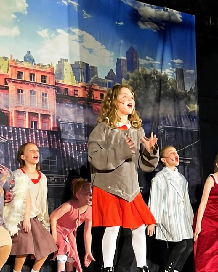 Full Annie Jr. cast performing on stage with a painted New York City backdrop