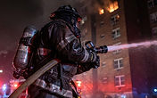 Firefighter in turnout gear working a hoseline at night outside a multi-story apartment building with visible smoke and glow from upper floors
