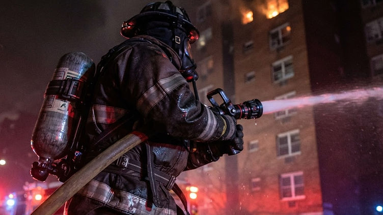Firefighter in turnout gear working a hoseline at night outside a multi-story apartment building with visible smoke and glow from upper floors