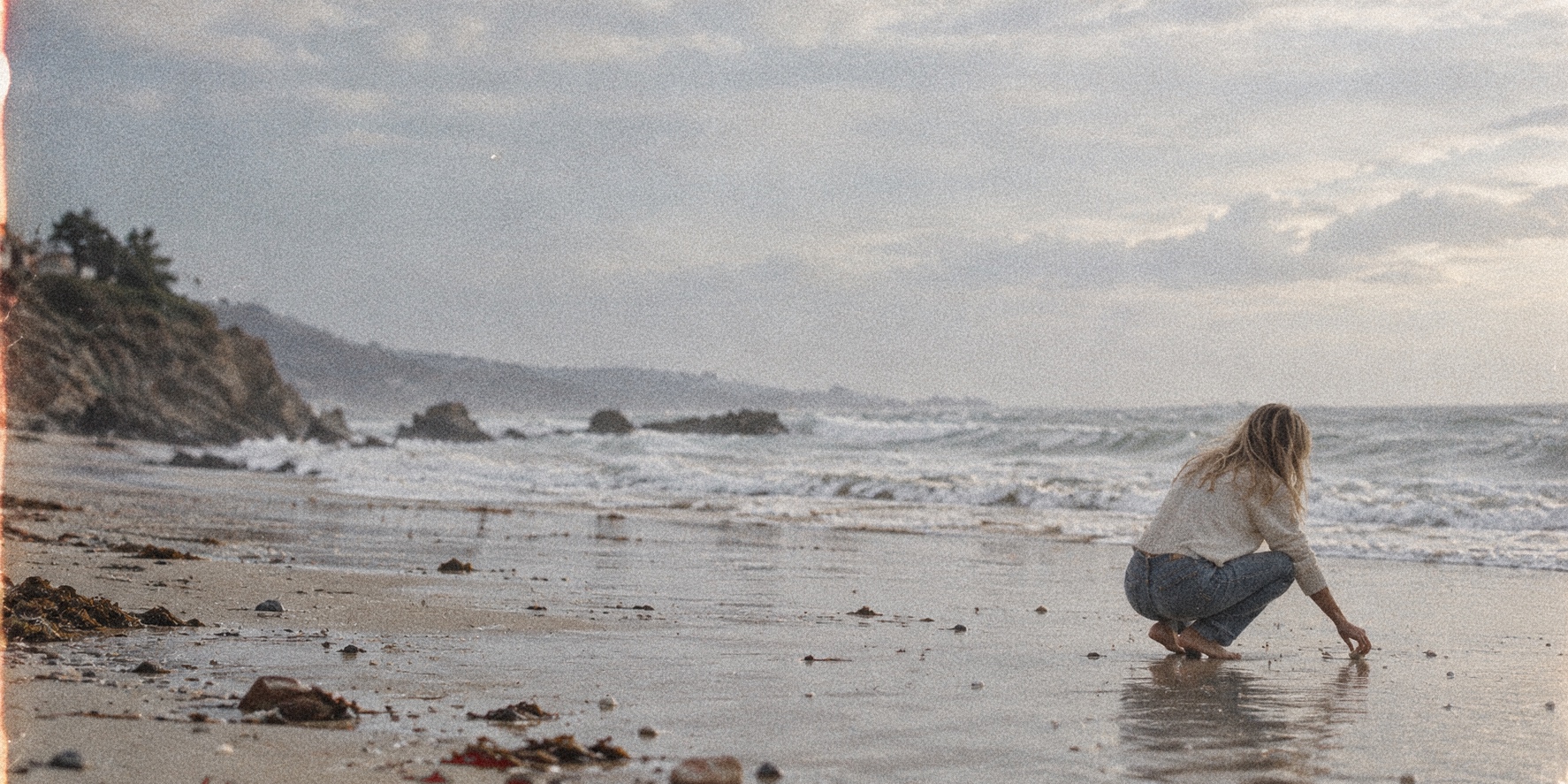 A woman crouching at the shoreline, reaching for shells in the wet sand at golden hour