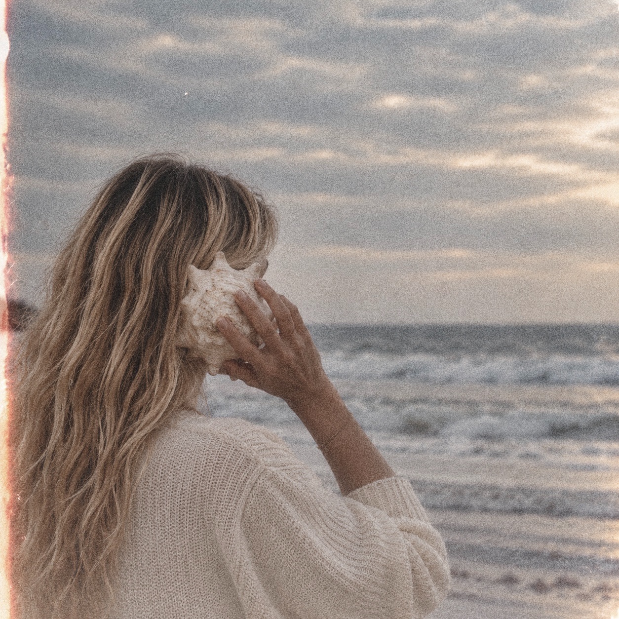 A woman holding a large seashell to her ear as waves roll in at golden hour
