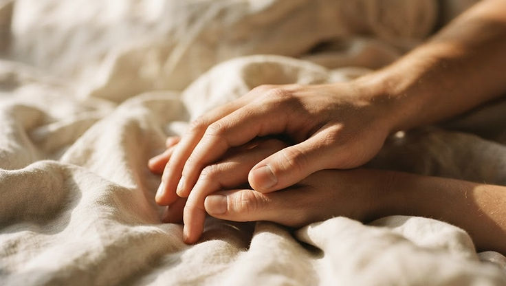 Two people's hands interlaced, resting together on soft bedding