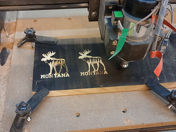 CNC router carving two moose and Montana designs into dark stock clamped to the bed