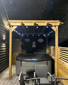 Private hot tub with its cover closed at night beneath a light-strung pergola at Lydden Bell Lodge