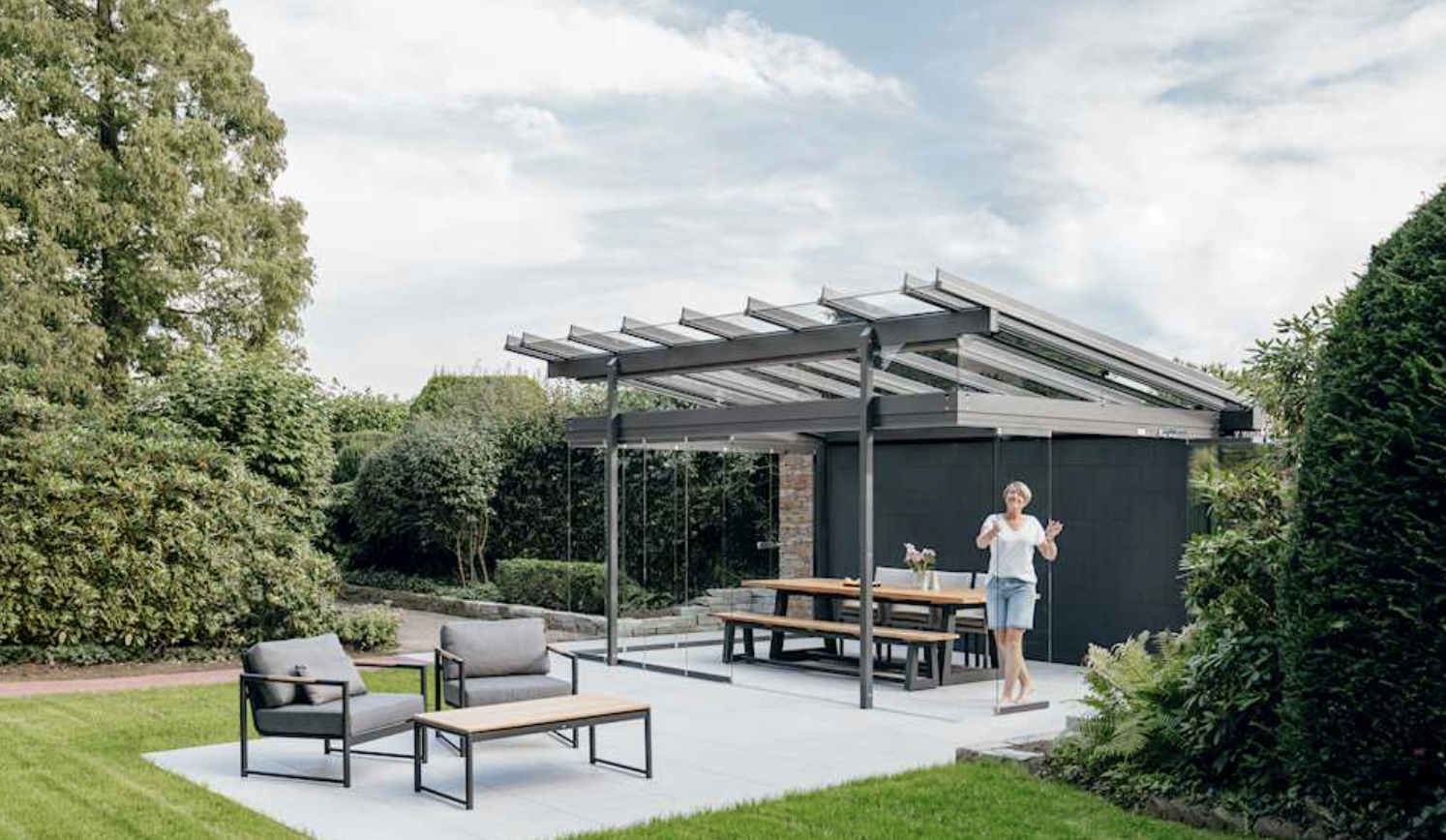 A canopy with sliding glass panels over a dining table on a paved patio against a brick house, the garden behind