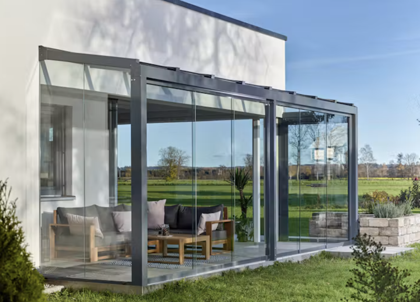A glass sliding wall closed across a patio lounge with a sofa, coffee table and planter, with fields beyond