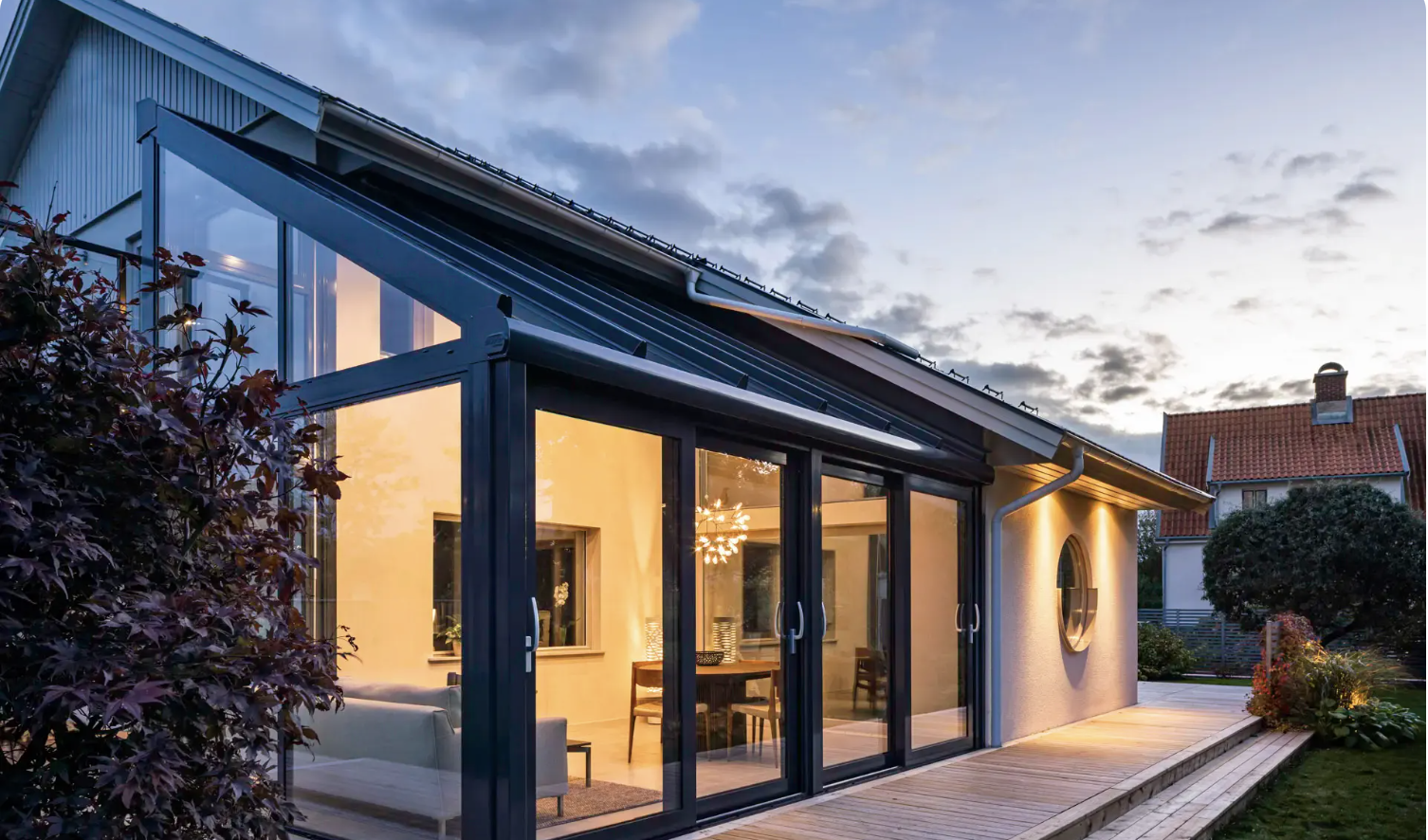A glass-roofed extension at twilight, its sliding doors open onto a timber deck with a maple beside the house