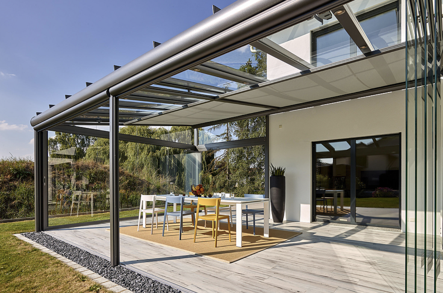 A glass canopy attached to a white house with its awning drawn out above a dining table on a timber deck