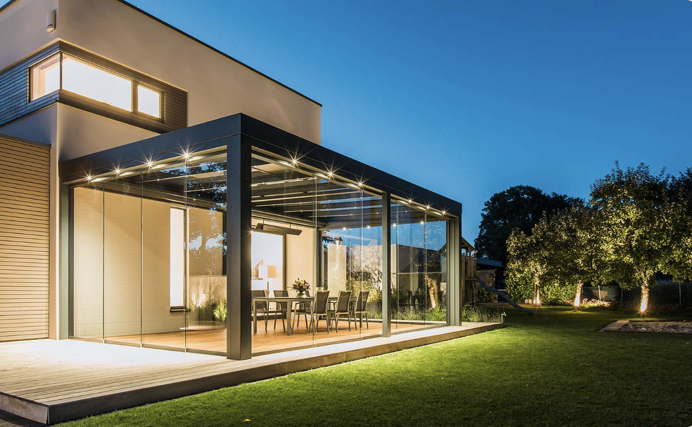 A dining table inside a dark-framed glass enclosure on a timber deck at dusk, opening onto a lawn
