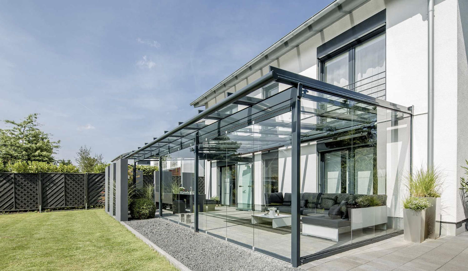 A lean-to glass canopy on dark posts sheltering a lounge set on a stone terrace beside a white house