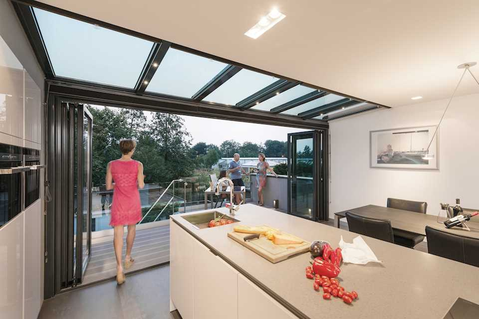 A white kitchen beneath a sloping glass roof, its folding doors open onto a balcony where guests stand at dusk