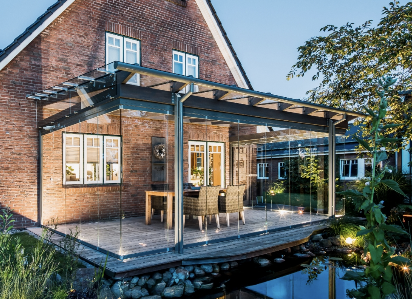 A glass-enclosed canopy over a dining deck against a red brick house, looking out across the garden pond