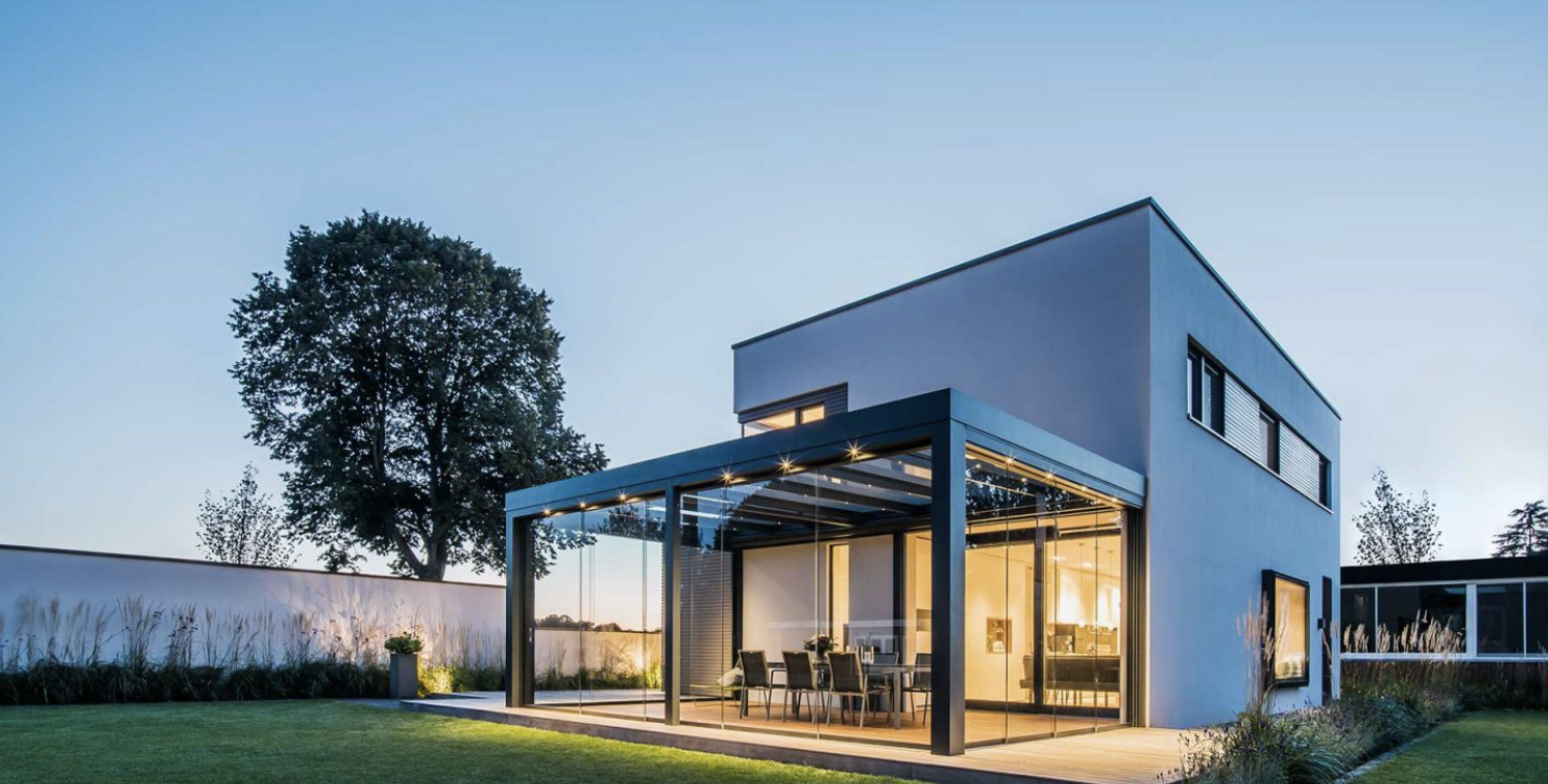 A glass canopy closed in at the rear of a two-storey house at twilight, its roof lights washing the glass above a lit deck