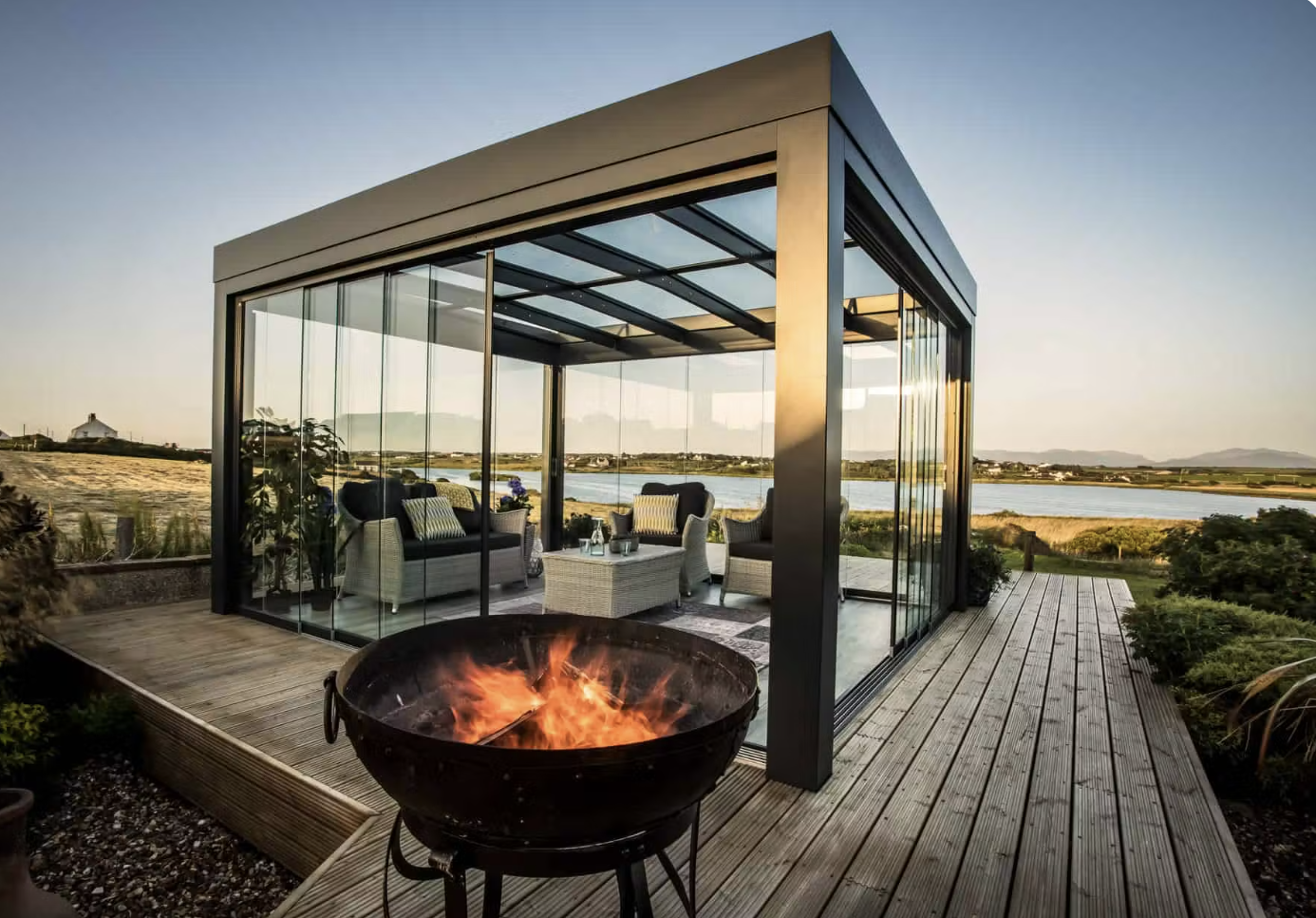 A free-standing glass canopy pavilion on a timber deck beside a lake, wicker lounge furniture inside and a fire bowl burning in front