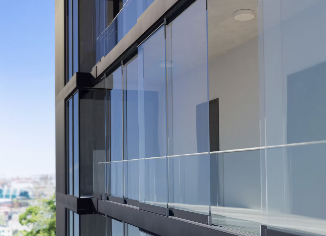 Frameless glass balcony glazing across a multi-storey facade, with city trees reflected in the panes