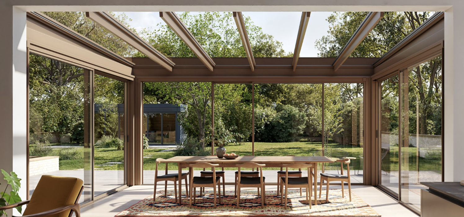 A long dining table beneath a bronze-framed glass roof, with the garden seen through the sliding doors in daylight