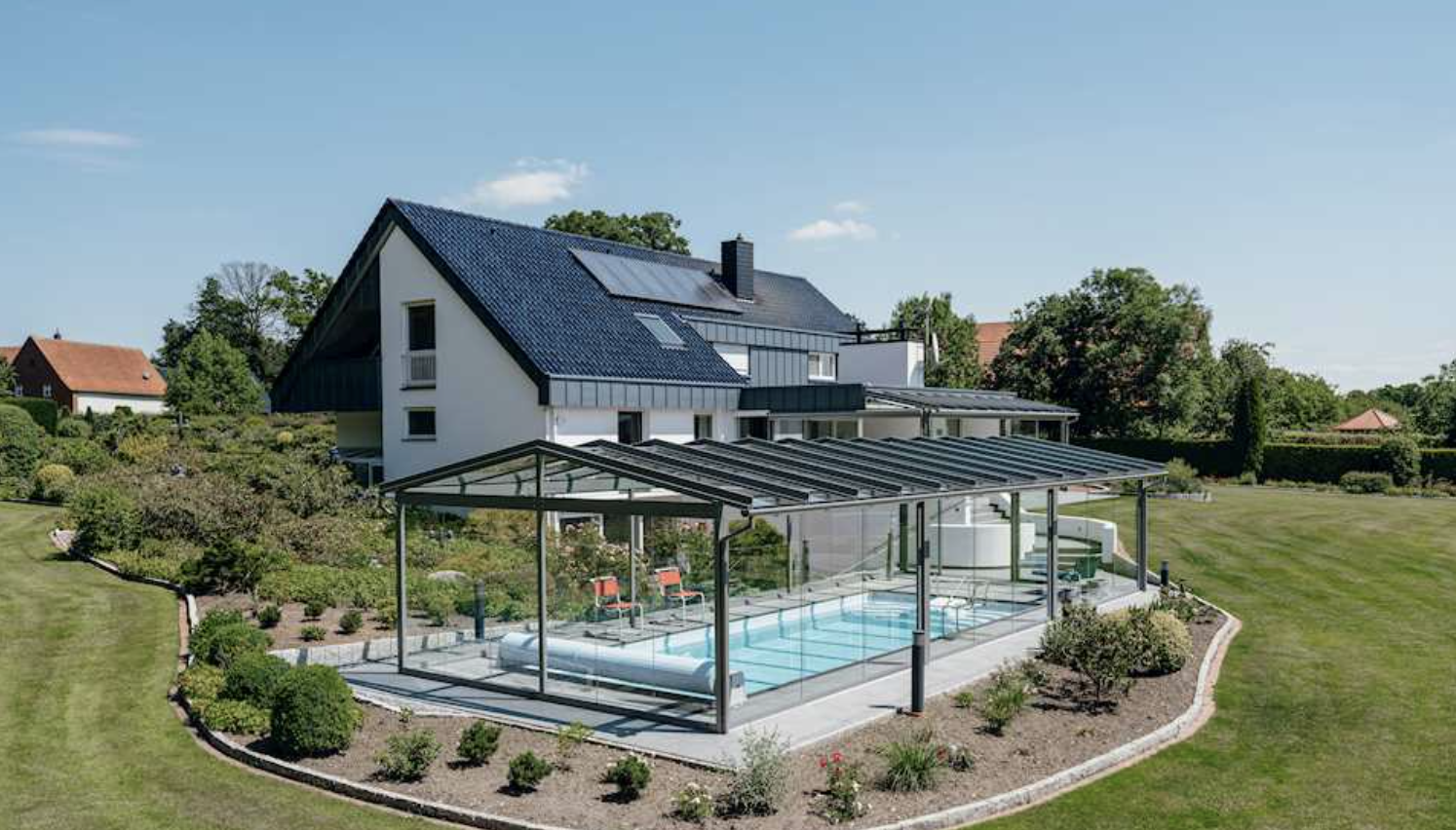 A dark-framed glass canopy over a swimming pool on a paved terrace beside a white house, with lawns and planting beyond