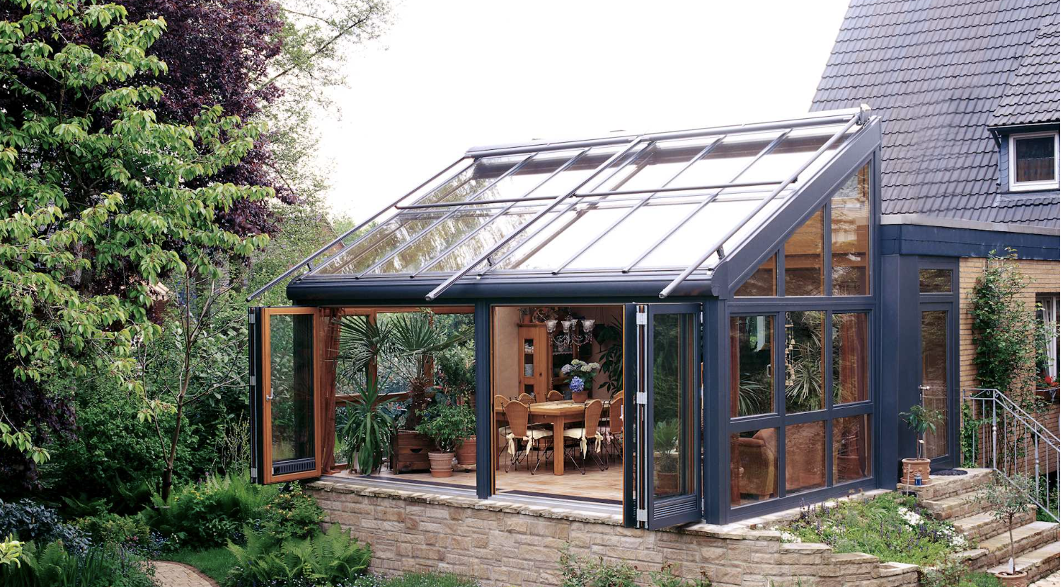 A wintergarden on a stone plinth against a brick house, its bifold doors open onto a tiered terrace with trees behind