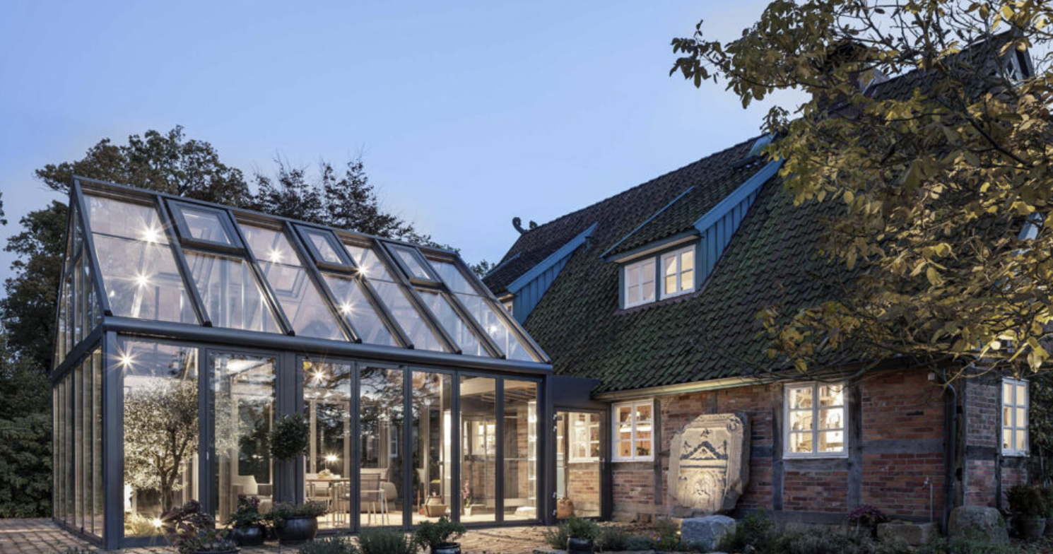 A dark-framed conservatory against a brick house at dusk, its glazed monopitch roof lit from within