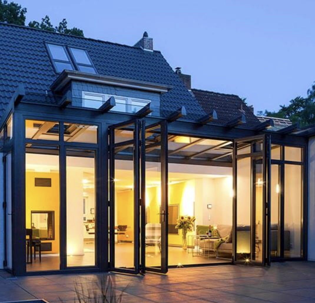 A black-framed wintergarden across the rear of a house at dusk, its folding doors open onto a stone patio with the living room lit inside