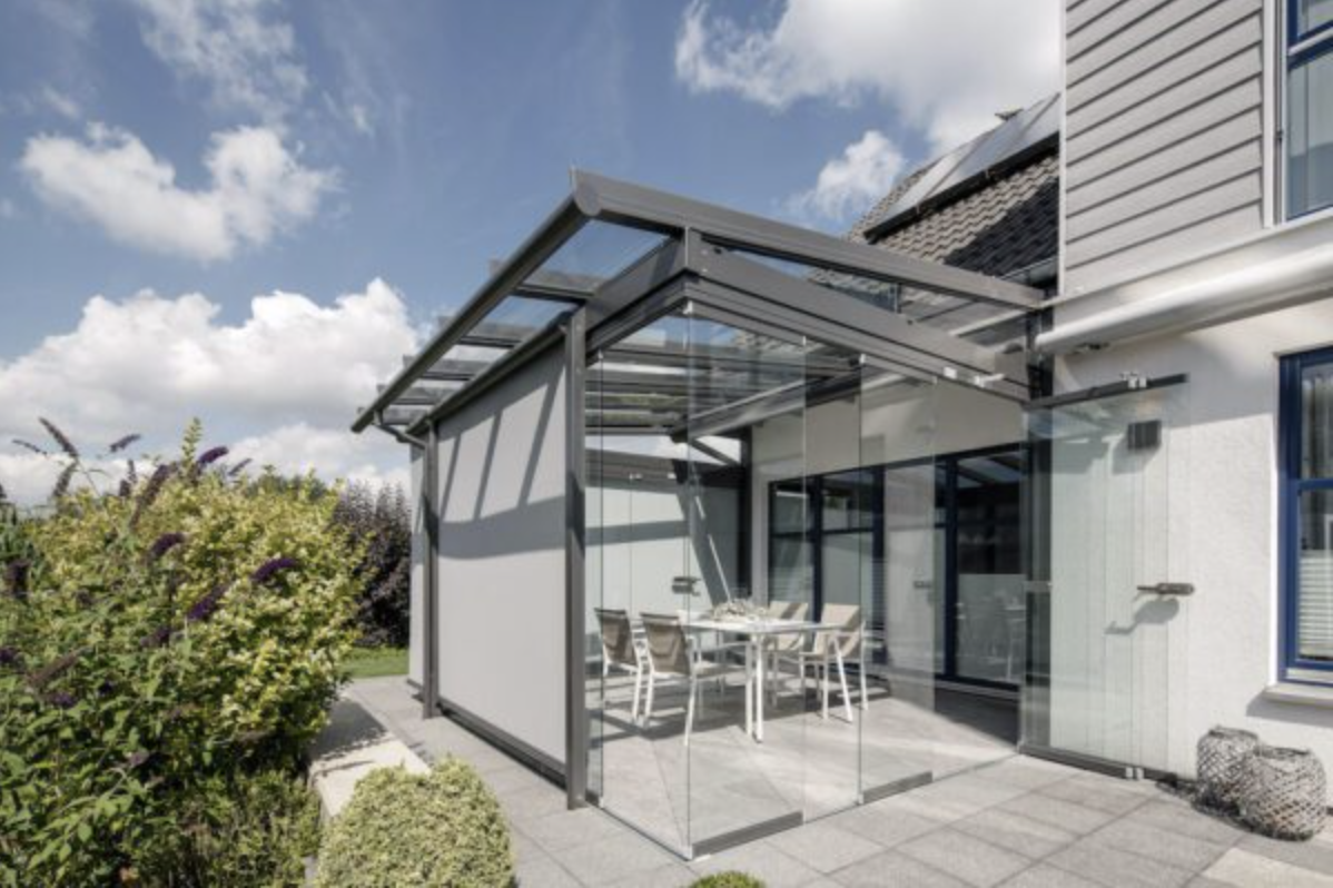 A glass canopy with a retractable side screen above a white dining set on a tiled patio, a flowering shrub beside it