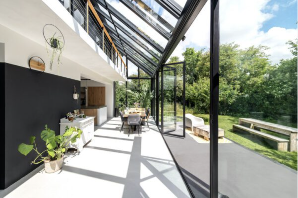 A dining table inside a black-framed wintergarden, its bifold doors open to a patio and garden in daylight