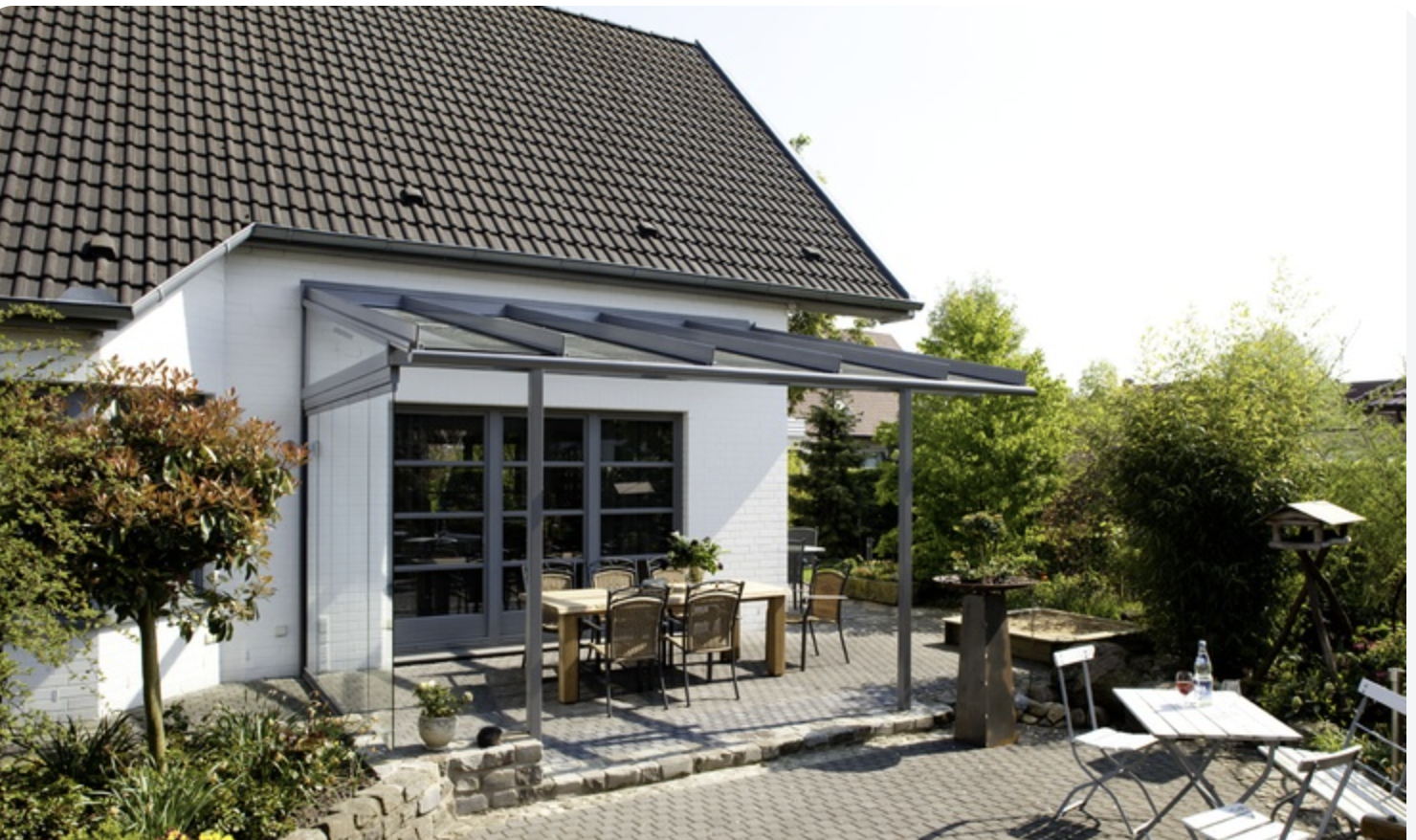 A sloping glass canopy on dark frames over a timber dining table, attached to a house with a pitched roof