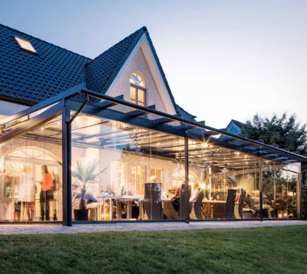 A glass veranda running across the rear of a gabled house at dusk, its dining table lit from within